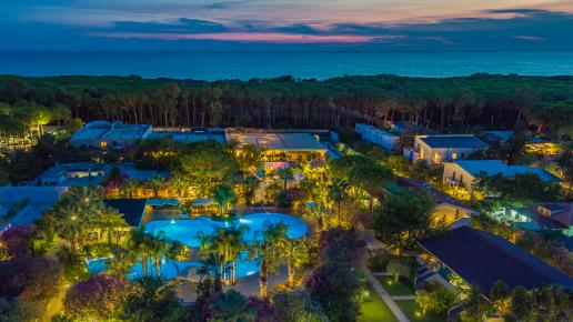 Aerial view of the resort surrounded by lush greenery, with an illuminated swimming pool and the sea in the background at sunset.
