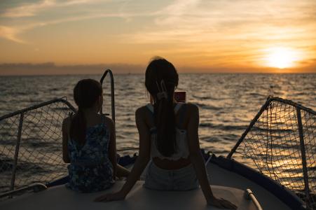Two girls on a boat admire the sunset over the sea, bathed in the golden light of the sun on the horizon.