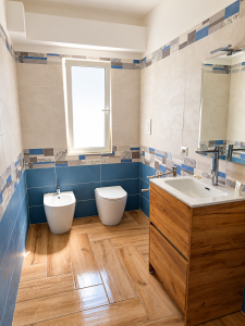 Modernes Badezimmer mit beigefarbenen und blauen Fliesen, Waschtischunterschrank aus Holz, weißen Sanitärobjekten und Holzoptik-Boden.
